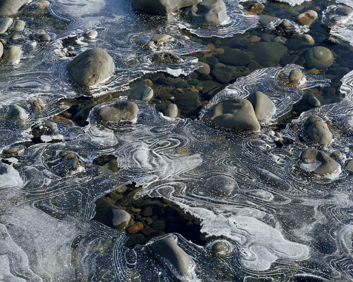 Photograph of a river's surface: eddies swirl in concentric lines as rocks peek to the surface. The color and texture of the sky and clouds above are hinted at in reflections on the water