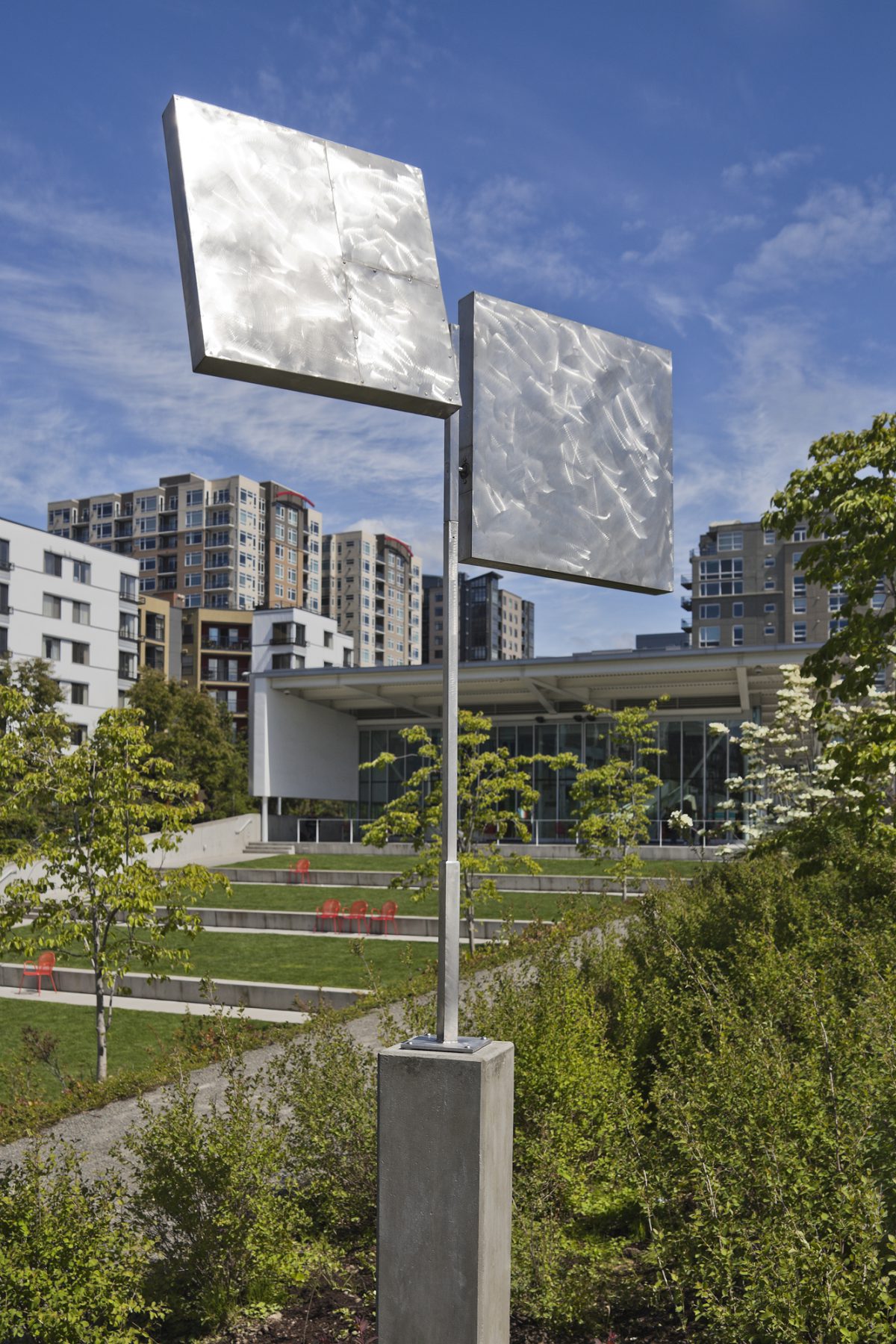 Vertical photo of a stainless steel sculpture, in the background is the Olympic Sculpture Park's Valley. A pole supports two planes that are burnished to reflect the sun's light in varying ways. The sculpture is elevated by a concrete plinth. Foliage dances along the lower half of the photo, while blue skies dotted with clouds inhabit the top.