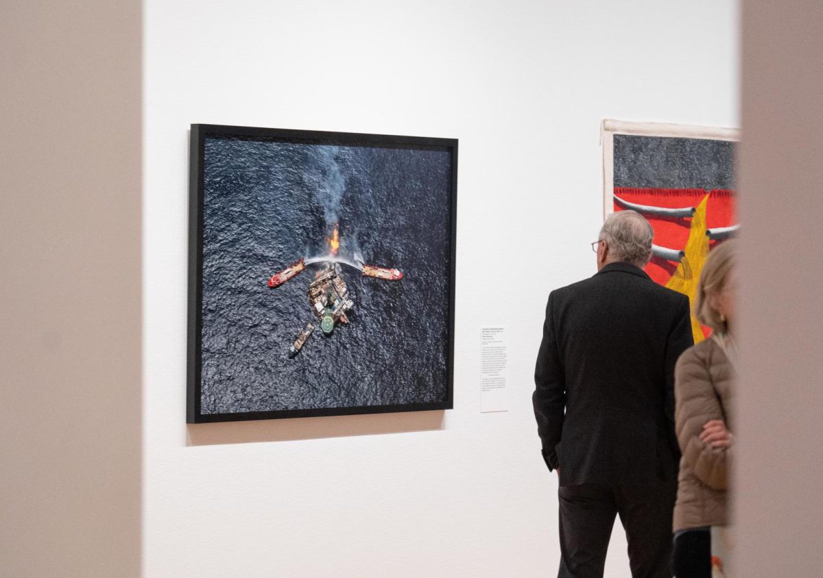 Gallery visitors looking at the artwork: This image is of the Deepwater Horizon oil spill in the Gulf of Mexico. The colors of the red emergency vehicles, the orange flare of the well flame, and the arc of water sprays appear minuscule against the backdrop of a blackening sea. 