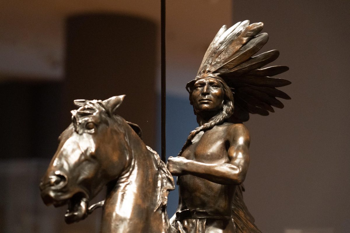 Photograph of the upper portion of a bronze sculpture of a Native American mounted on a horse. The man depicted wears a large feathered headdress and long braids draped over his bare chest. He holds the reigns in his left hand and an erect spear in his right, while looking stoically in the distance.
