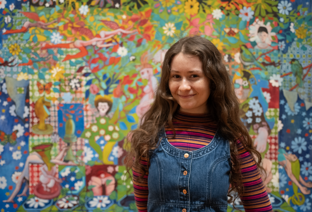 A young woman with long brown curly hair stands in front of a vibrant painting.