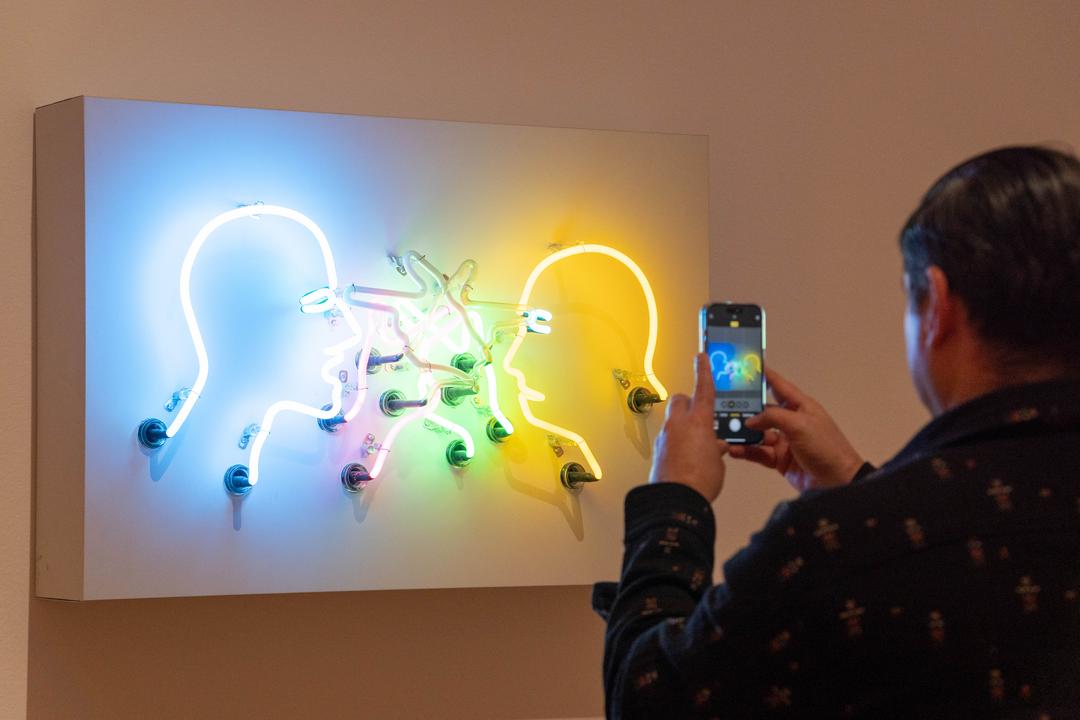 A man holds his phone in front of a neon art piece and takes a picture