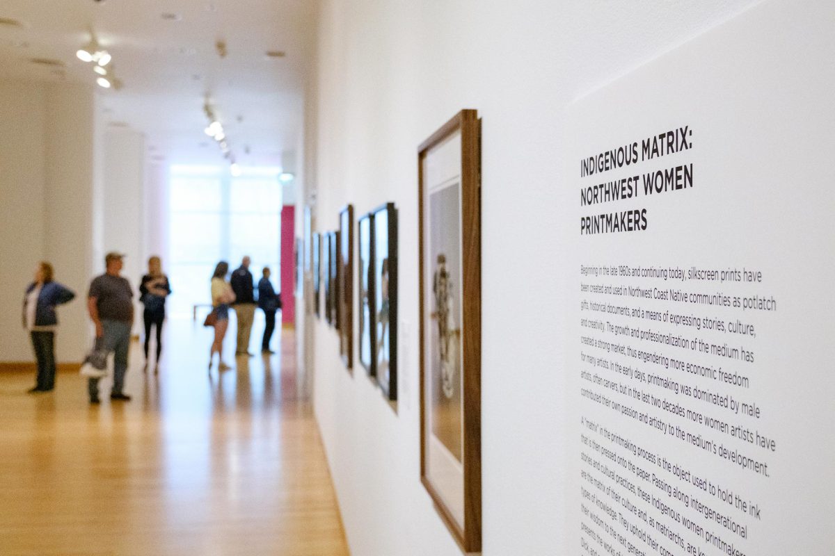 Photograph of a long hallway, the wall is lined with framed screenprints. Viewers stand in the distance on the left. To the right is the exhibition's interpretive text