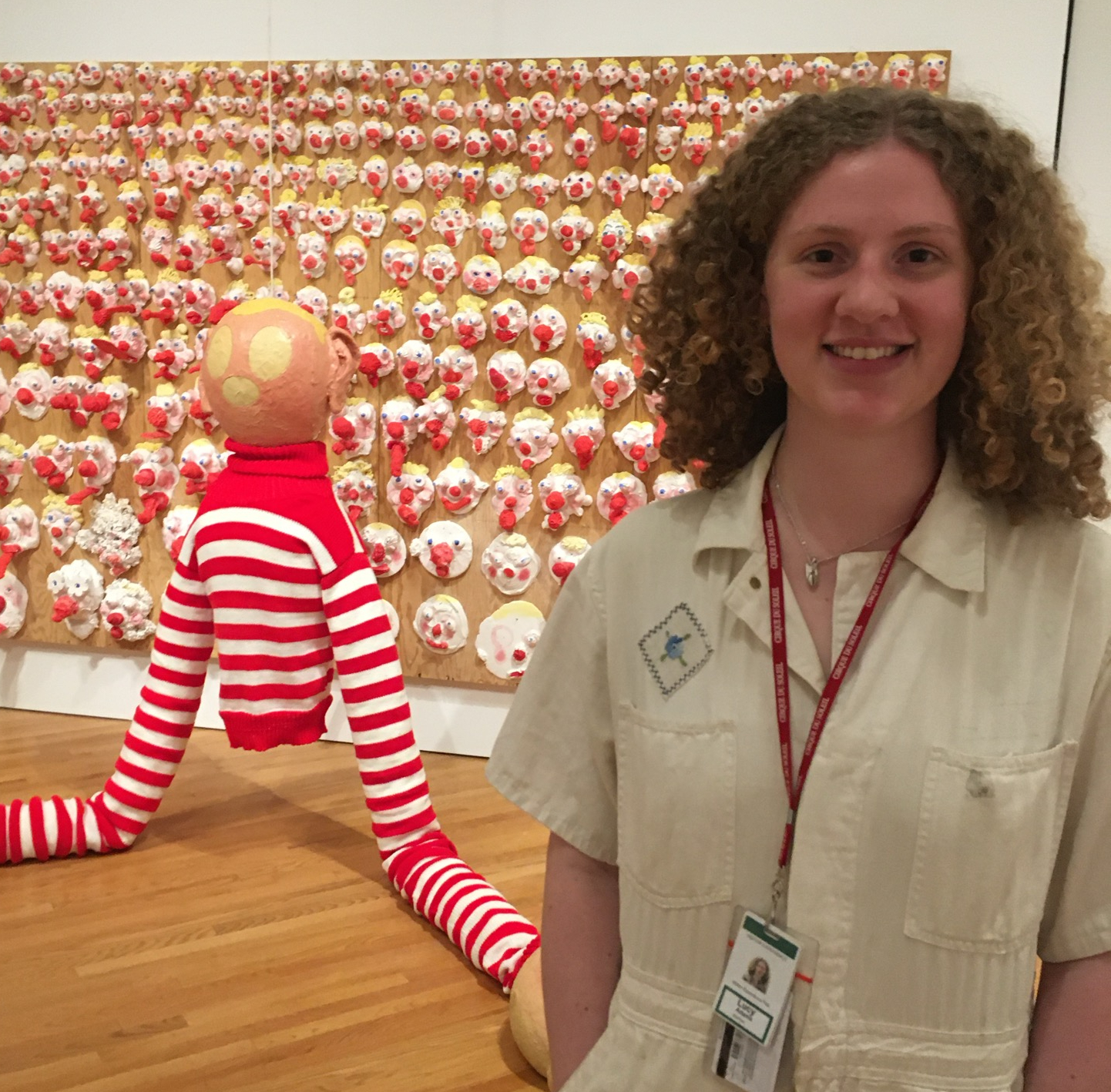 A woman with curly medium length hair stands in front of an art installation featuring clown papier mache.