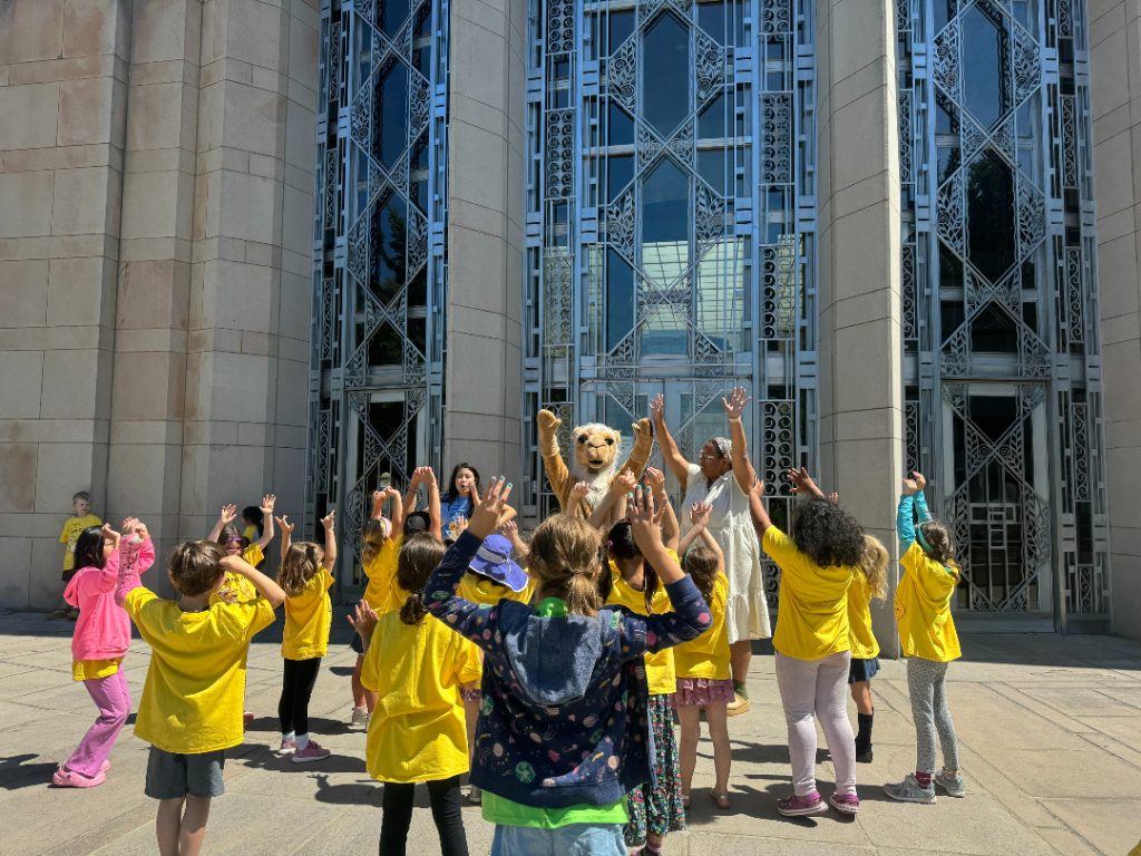 A group of children cheer with a camel mascot in front of an Art Deco building.