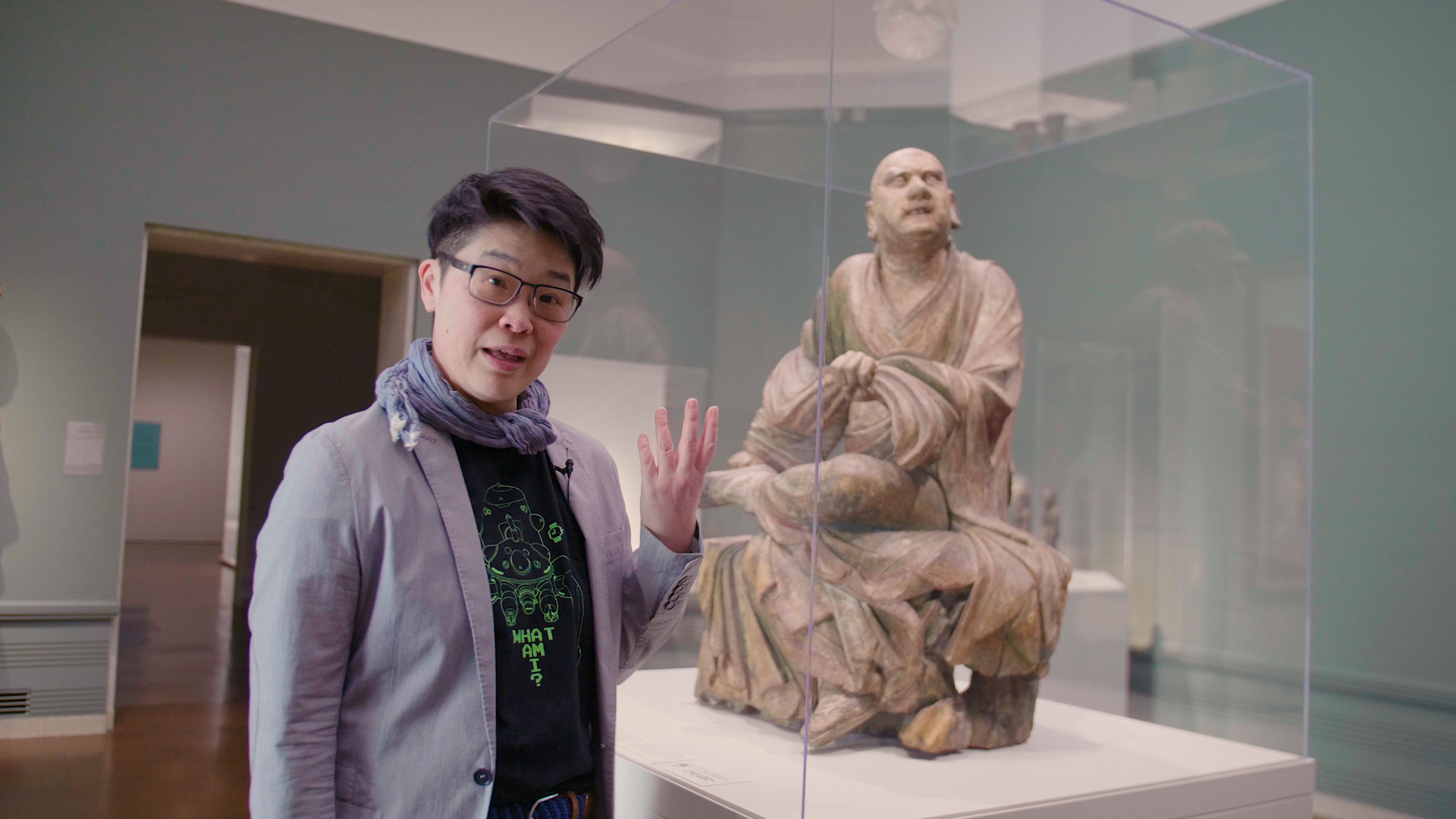 An art curator gestures to a large wooden Luohan sculpture behind her.